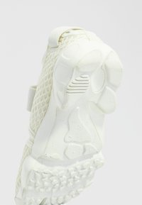 Shoe in off-white mesh and rubber, featuring a textured outsole with varied patterns, reinforced toe, and a soft fabric strap.