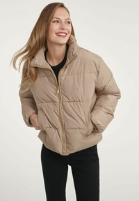 Beżowa kurtka puchowa z wysokim kołnierzem i pikowanym wzorem, z zamkiem błyskawicznym z przodu i bocznymi kieszeniami. Złote akcenty na zamku.