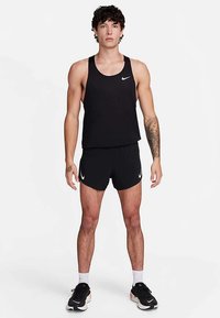 Top deportivo negro sin mangas y pantalones cortos con el logo de la marca Nike en blanco, diseño ajustado, tejido suave y ribete contrastante. El modelo está de pie mirando de frente.