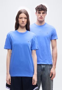 Νεαρή γυναίκα και άνδρας στέκονται δίπλα-δίπλα, και οι δύο φορούν ανοιχτό μπλε μπλουζάκια με μικρά λογότυπα, με ουδέτερες εκφράσεις, απλό λευκό φόντο.