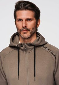 Sudadera con cremallera en tono beige, confeccionada con tela suave, que cuenta con una capucha con cordón, detalles en negro y texto bordado en el hombro.