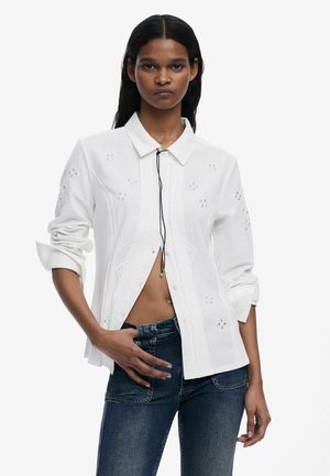 Camicia bianca con bottoni, caratterizzata da cuciture decorative, colletto e maniche arrotolate, abbinata a jeans in denim scuri.