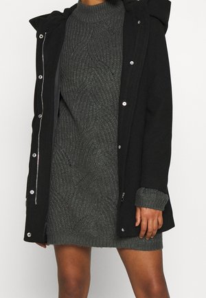 Femme portant une robe tricotée gris foncé à manches longues et un manteau noir à capuche avec des boutons-pression partiellement ouverts.