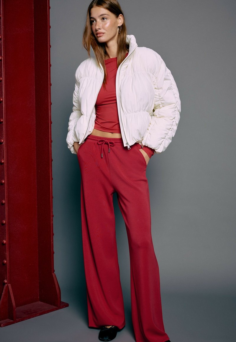 Femme aux cheveux bruns raides porte une doudoune blanche sur un crop top rouge et un pantalon large rouge, debout devant un fond gris et rouge.