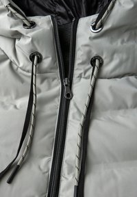 Ljusgrå pufferjacka med hög krage, svart dragkedja, svart foder och justerbara dragsnoddar. Har metallöglor och strukturerade snören.