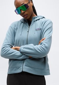 Άτομο που φοράει γαλάζιο φλις σακάκι Patagonia και μεγάλα ανακλαστικά γυαλιά ηλίου με τα χέρια σταυρωμένα μπροστά σε λευκό φόντο.