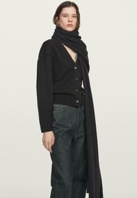 Czarny dzianinowy kardigan z głębokim dekoltem w serek i zapięciem na guziki, zestawiony z długim czarnym szalikiem. Noszony z ciemnymi dżinsami, wykonany z teksturowanej tkaniny.