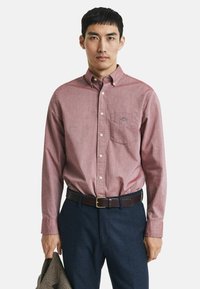 Hemd zum Zuschneiden mit rot-weißem Karomuster, Baumwollstoff, lange Ärmel, Vordertasche, getragen mit dunkler Hose und braunem Gürtel.