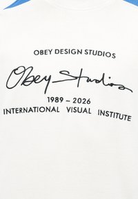 Λευκό ύφασμα με μαύρο κείμενο που γράφει "OBEY DESIGN STUDIOS", μια καλλιγραφική υπογραφή "Obey Studios" και "1989 - 2026 INTERNATIONAL VISUAL INSTITUTE."