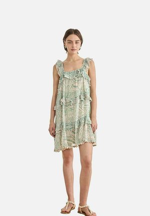 Mujer con un vestido sin mangas de patrón verde claro y beige con volantes, de pie descalza contra un fondo blanco liso.