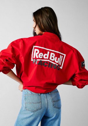Červená vetrovka v jasnej červenej farbe s bielym a čiernym logom "Red Bull RACING" na chrbte, doplnená modrými rifľami.