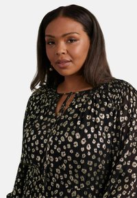 Svart klänning med metallisk guldleopardtryck, rund halsringning och knytöppning. Har långa ärmar och drapering vid axlarna.