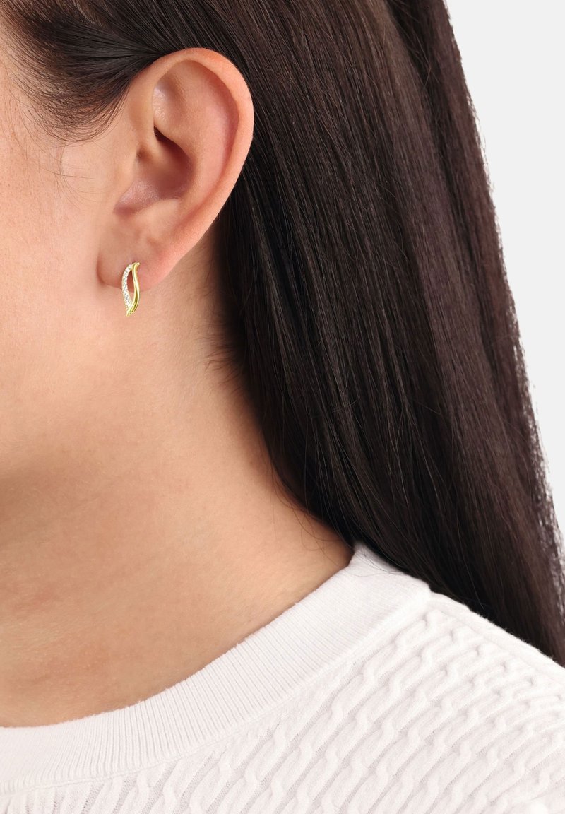 Örhänge i guldton med bladform som har en strukturerad yta och en rad små klara stenar. Bärs i ett öra, med hår delvis synligt.