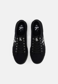 Zapatillas de lona negras con punta redonda, suela de goma, cordones negros y ojetes plateados. Presenta el logo en el lateral y una etiqueta en la plantilla.