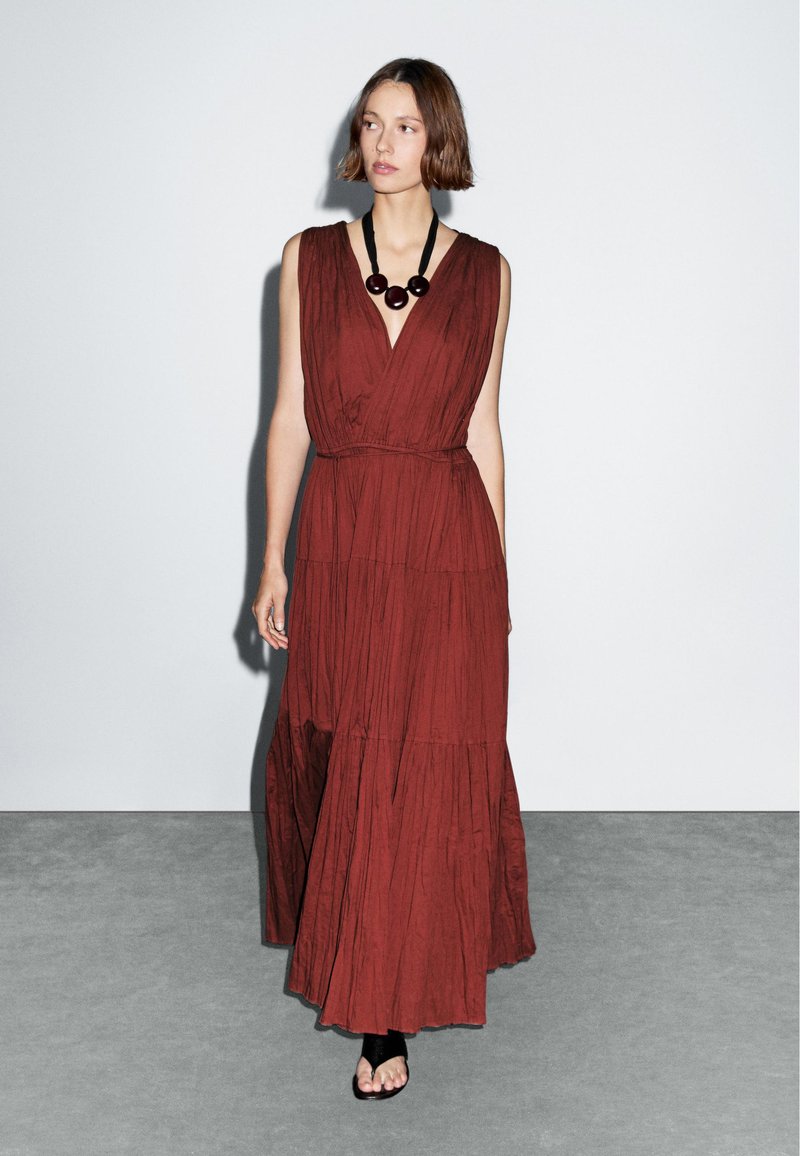 Robe maxi rouge à volants avec décolleté en V profond et ceinture à la taille, fabriquée en tissu texturé. Assortie à un collier en perles noir et des sandales à lanières.