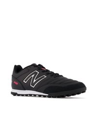 Black athletic shoe with synthetic upper, textured accents, white logo, small pink detail, and rubber studs for grip on the outsole.