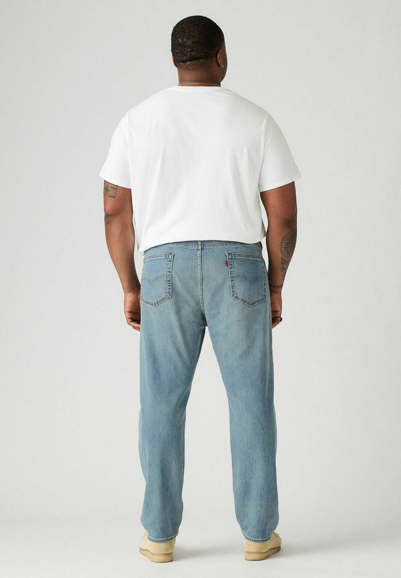 Jeans en denim bleu clair avec une coupe droite et deux poches arrière. Portés avec un t-shirt blanc uni à manches courtes et des chaussures beiges.