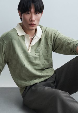 Jeune homme assis portant un polo en maille à carreaux vert pâle avec un petit logo et un collier de perles, pantalon gris, fond neutre.