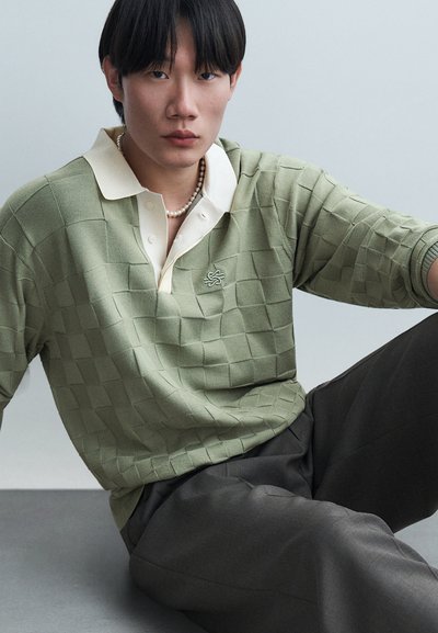 Junger Mann sitzt, trägt ein hellgrünes kariertes Strick-Poloshirt mit kleinem Logo und eine Perlenkette, graue Hose, neutraler Hintergrund.