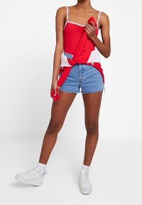 Rotes ärmelloses Oberteil mit weißen Akzenten, um die Taille geknotet, kombiniert mit Jeansshorts. Das Modell trägt weiße Sneakers und Knöchelsocken.