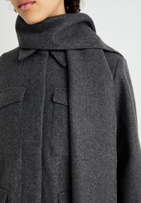 Cappotto in lana grigia con un ampio colletto a sciarpa, dotato di due tasche frontali e una finitura morbida e strutturata, progettato per il calore e il comfort.