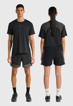 Mann, der nach vorne schaut, und Frau, die nach hinten schaut, tragen schwarze Sportshirts und Shorts und stehen vor einem schlichten hellen Hintergrund.