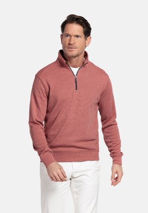 Man wearing rust-colored quarter-zip pullover and white pants, standing against a plain light background.