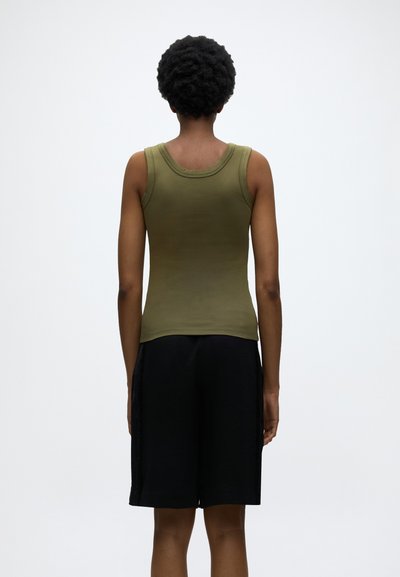 Άτομο με κοντά σγουρά μαλλιά που φοράει ελαιοπράσινο αμάνικο τοπ και μαύρο σορτς μέχρι το γόνατο, εμφανιζόμενο από πίσω.