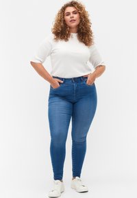 Jeans ajustados azules de cintura alta, con passadores y bolsillos delanteros, combinados con una camiseta de manga corta blanca con textura acanalada y zapatillas blancas.