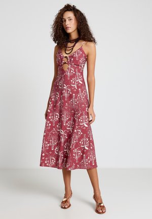 Woman with curly hair wearing a sleeveless, patterned red midi dress with front cutouts and brown heeled sandals, standing on a plain background.