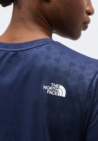 Κοντινό πλάνο στο άνω μέρος της πλάτης ενός ατόμου που φοράει μπλε σκούρο πουκάμισο με λεπτό μοτίβο και λογότυπο The North Face σε λευκό χρώμα.