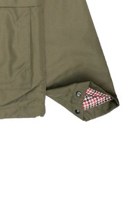 Manga de chaqueta verde oliva con un forro de patrón de pata de gallo, que cuenta con un cierre de botón a presión y un bolsillo de parche en el brazo.