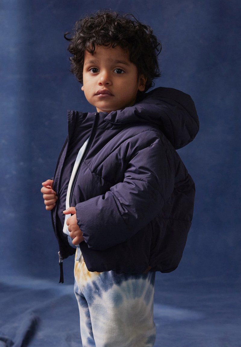 Marinblå puffjacka med huva och zip, bärs över en vit topp. Barnet matchar den med tie-dye sweatpants med blå och guld mönster.