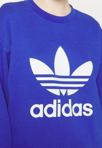Blå sweatshirt med stort vitt Adidas trefoil-logotyp och text framtill, buren av en person som visas från halsen till mitten av torso.