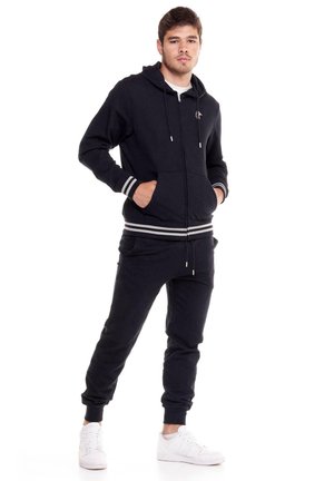 Un hombre luce una sudadera negra con capucha y cremallera, a juego con unos pantalones de chándal con rayas blancas. Está de pie con las manos en los bolsillos sobre un fondo blanco.
