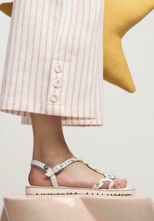 Sandalia blanca con tiras cruzadas y suela texturizada, llevada en un pie al lado de pantalones a rayas rosa claro y blancos con detalles de botones.