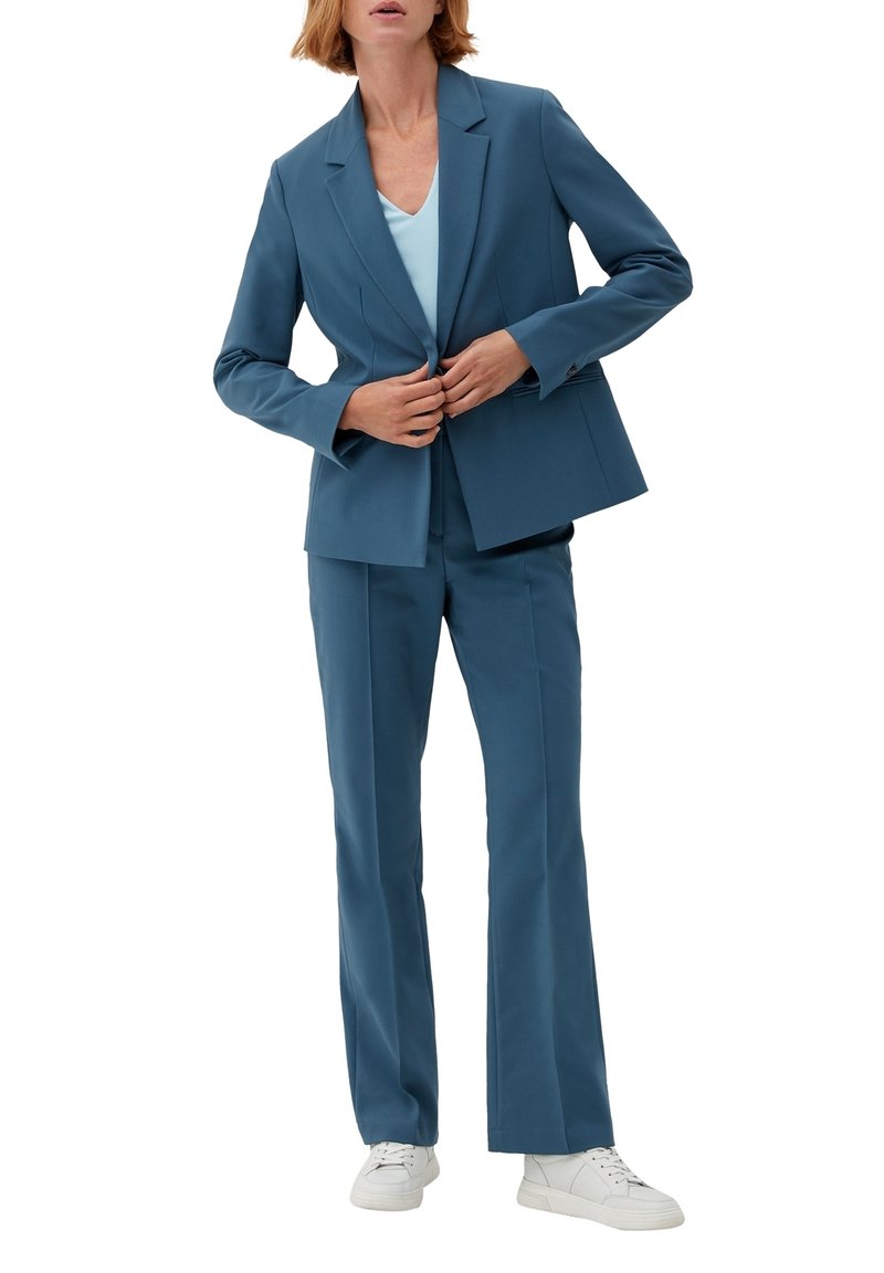 Femme boutonnant un blazer bleu porté avec un pantalon assorti, un haut bleu clair et des baskets blanches sur fond blanc.
