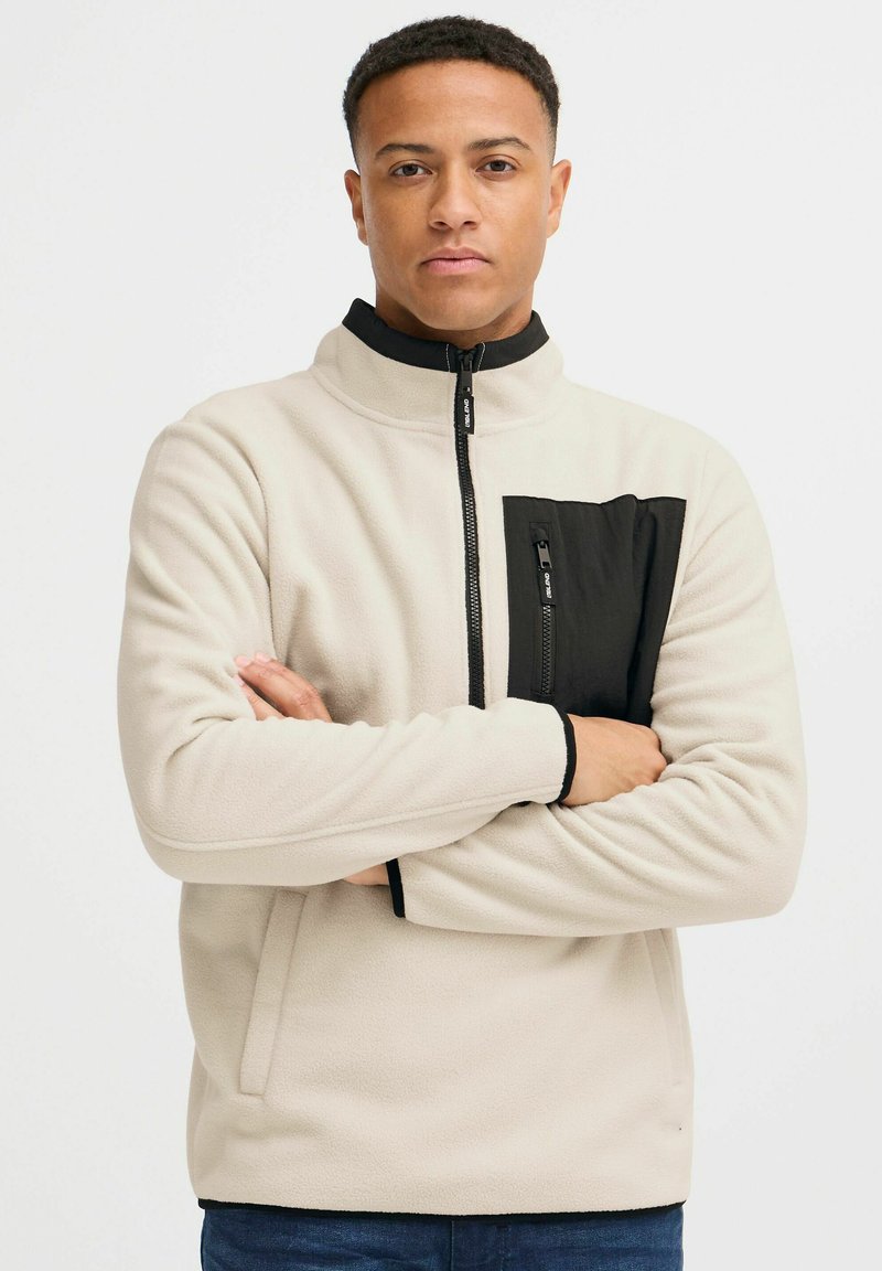 Chaqueta de polar beige con detalles en negro, cuenta con un bolsillo en el pecho con cremallera, cuello alzado y puños negros contrastantes.