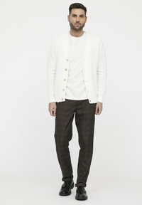 Weißer Strickcardigan mit Holzknöpfen, getragen über einem weißen Hemd, kombiniert mit braunen karrierten Hosen und schwarzen Schuhen. Einfaches Design.