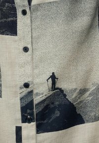 Camicia a maniche corte con bottoni realizzata in tessuto leggero, con sfondo beige e stampe grafiche scure di un escursionista e forme astratte.