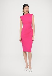 Dopasowana sukienka do kolan w jasnoróżowym kolorze, z wysokim dekoltem, krótkimi rękawkami i marszczeniem w talii. Połączona z białymi szpilkami.