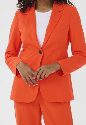 Femme portant une veste de costume ajustée orange vif et un pantalon assorti sur un haut blanc, les mains doucement croisées à la taille.