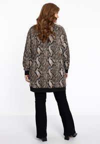 Lange, oversized cardigan met een slangprint in bruin, zwart en crème, afgewerkt met geribde manchetten en zoom, gecombineerd met zwarte flared broek.