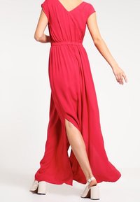 Robe longue et fluide en tissu rose vif avec un décolleté en V et des manches courtes. Présente une taille cintrée et des fentes sur les côtés, associée à des talons blocs blancs.