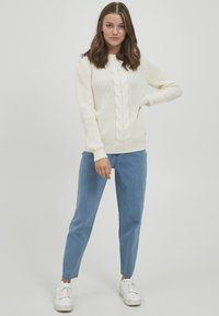 Kremfarget kabelstrikket genser med lange ermer, ribbestrikkede mansjetter og kant. Kombinert med lyseblå avsmalnende jeans og hvite joggesko. Enkelt bakgrunn.