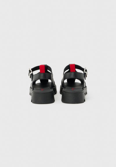 Μαύρα σανδάλια με χοντρό τακούνι, ανοιχτή πλάτη, κόκκινη ταινία τραβήγματος και ρυθμιζόμενα λουριά με ασημένια αγκράφα, εμφανίζονται από πίσω σε ανοιχτό φόντο.