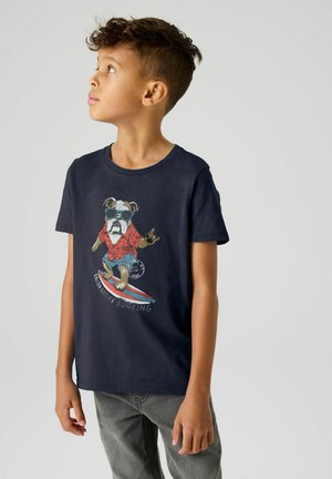 Jonge jongen in een donker T-shirt met een afbeelding van een surfende hond die een zonnebril en een rood shirt draagt, staand tegen een grijze achtergrond.