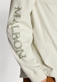 Long-sleeve crewneck shirt in light beige cotton with green graphic text and ornate design detailing on the left sleeve.