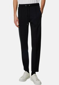 Svarta sweatpants med dragsko i midjan, tapered fit och fickor med dragkedja; kombinerat med vita sneakers och en ljusgrå t-shirt.