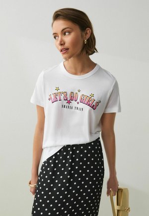 Kvinde iført hvid "Let's Go Girls Shania Twain" t-shirt og sort nederdel med polkaprikker, holder en beige håndtaske og kigger til siden.